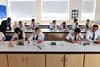 High school students wearing school uniform  and safety glasses do written work in a school lab. The chemistry teacher is helping some of the students.