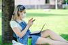 A photo of a student with a face mask studying on a laptop and mobile phone in a park