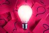 Bright lightbulb on a background of pink paper squares with questions marks written on them