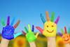 Four hands held up against a blurred, spring feild background, each are painted different bring colours, fingers spread and with smiley faces painted on to the palm
