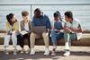 A group of four young people sit outdoors with notebooks while one person shows content on a laptop, all gathered on a wooden bench