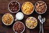 A variety of different breakfast cereals in bowls on a wooden surface, with a glass jug of milk and spoons at the side