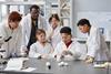 Group of children in lab coats and safety glasses doing a practical with their teacher looking on