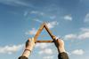 two hands holding a wooden triangle up to a blue sky with clouds in it