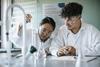 A teenage girl and boy doing an experiment in a school chemistry lab. They are wearing lab coats and safety goggles. The girl sis using a plastic pipette while the boy watches. Behind them is a poster of Chemistry Week 2025.