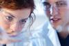 Two students wearing safety goggles and lab coats closely observe a reaction