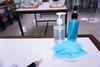 An image showing a medical mask, bottles of hand sanitzer and pencils on a desk in a school classroom.