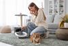 A woman holds her nose as she uses a scoop to remove used litter from a cat litter tray. A ginger cat sits nearby.