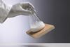 A photo of a hand in a latex glove holding a conical flask of white powder with a damp piece of wood stuck to its base