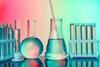 A selection of glass laboratory equipment, including test tubes in a test tube holder, a measuring cylinder, a conical flask and a round-bottom flask, against a colourful background