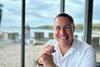 Adam Savin is smiling at the camera, wearing a white shirt. The sea is in the background.