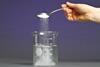 A hand pouring a spoon of sugar into a beaker of water