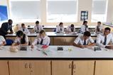 High school students wearing school uniform  and safety glasses do written work in a school lab. The chemistry teacher is helping some of the students.