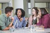 High school students in a lab discussing chemistry. They are wearing safety goggle and there is lab equipment set up around them.
