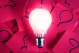 Bright lightbulb on a background of pink paper squares with questions marks written on them