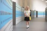 An image showing a shy female student wearing school uniform with a yellow satchel walking with her head down on a corridor lined by blue and grey lockers