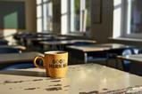 A colourful mug which says Good Morning with steaming coffee on a teacher's desk at the front of an empty classroom before the school day begins.