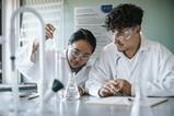 A teenage girl and boy doing an experiment in a school chemistry lab. They are wearing lab coats and safety goggles. The girl sis using a plastic pipette while the boy watches. Behind them is a poster of Chemistry Week 2025.