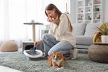 A woman holds her nose as she uses a scoop to remove used litter from a cat litter tray. A ginger cat sits nearby.