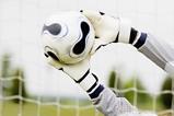 A football goalkeeper wearing protective gloves stretching to block a football from going into the goal