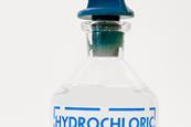 A close-up image of a glass bottle of dilute hydrochloric acid with a blue stopper