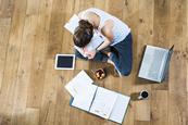 An image from above showing a girl sat cross legged, revising for exams, surrounded by a laptop, tablet, notes, coffee and snack