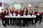 A group of secondary school students standing around a table in a classroom. The students are the front of the group are holding tablets displaying the home page of the I'm a Scientist, Get me out of here website.