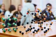 Models of different types of chemical bonding including diamond and salt on a table in a classroom. Behind a teacher speaks to the high school students.