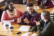 Three students sitting at a table with a tablet, pens, paper, having a discussion