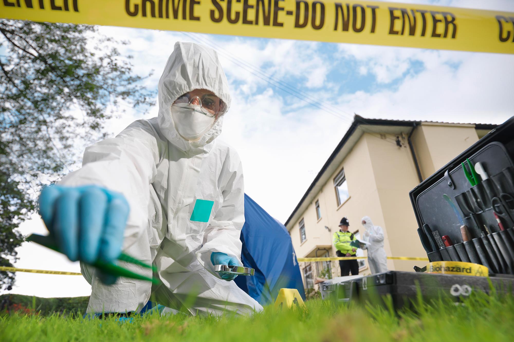 Forensic investigator in white hazmat suit spraying luminol at a residential outdoor crime scene near a house and police tape
