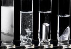 Four test tubes in a row with metals reacting in a clear liquid increasing in bubbles from left to right