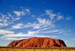 Uluru in Australia - a large orange-coloured rocky hill in a dry landscape with a cloudy blue sky