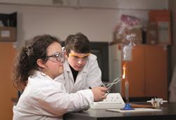 An image showing a female student burning magnesium on a Bunsen burner using tongs while a male classmate is watching the flame created. Both are wearing safety goggles