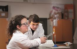 An image showing a female student burning magnesium on a Bunsen burner using tongs while a male classmate is watching the flame created. Both are wearing safety goggles