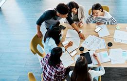 A photograph from above showing a group of university students discussing their work together, with phones, notebooks and documents in front of them