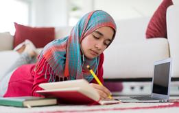 A female student wearing a hijab studies at home, using a notebook, pen and laptop