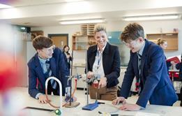 A photograph of two secondary school students and their teacher smiling while discussing an experiment in a chemistry lesson