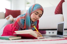 A female student wearing a hijab studies at home, using a notebook, pen and laptop