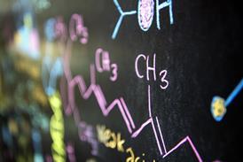 A close-up photograph of a chemical formula written in pink chalk on a blackboard, with other chemical structures drawn in different colours in the background