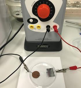 Photo showing set up for electroplating a coin at microscale. The positive terminal is connected to a paper clip holding zinc foil in a Petri dish with an electrolyte containing zinc ions. Negative terminal connects to paper clip holding copper coin.