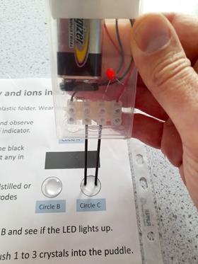 A hand holding two electrodes in a drop of water on a laminated sheet of paper. There is a red LED lit up.