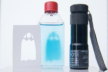 A torch, a stencil of a ghost and a glass bottle with a clear liquid that has the shape of a blue ghost within the liquid