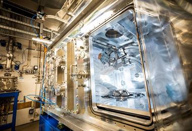 A technician in a white cleansuit makes adjustments inside a large piece of metal scientific equipment