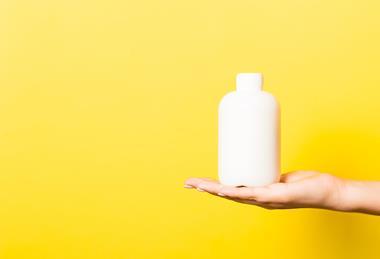A photograph showing an open hand holding a plain white shampoo bottle in front of a yellow background