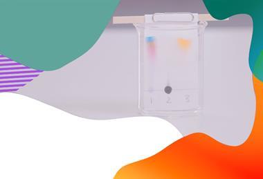 Chromatogram sitting in a beaker. The paper has a pencil line with three labels, marked as 1, 2 and 3. Sample 1 has separated into blue, orange and pink up the paper. Sample 2 is still a black circle of ink on the pencil line and sample 3 contains orange