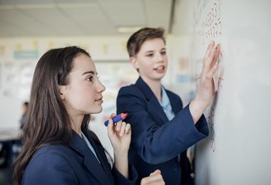 A photograph showing two students writing on a whiteboard in their classroom