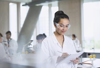 An image showing a female student writing notes in a notebook she's holding; she is wearing a lab coat and lab spectacles, and the background is a school science laboratory