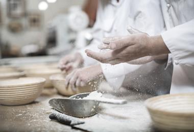 A photograph of a baker's hands covered in flour, as the baker prepares ceramic bowls for baking bread