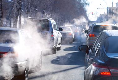 A photograph of queuing urban traffic, showing steam rising from car exhaust pipes