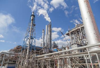 A fractional distillation tower as part of a large industrial plant with lots of pipes, scaffolding and chimneys giving off emissions.